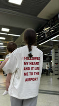 A person standing in an airport terminal wearing a white oversized T-shirt from the back, which features the red text 'I FOLLOWED MY HEART AND IT LED ME TO THE AIRPORT'. The person is holding a baby on their shoulder.
