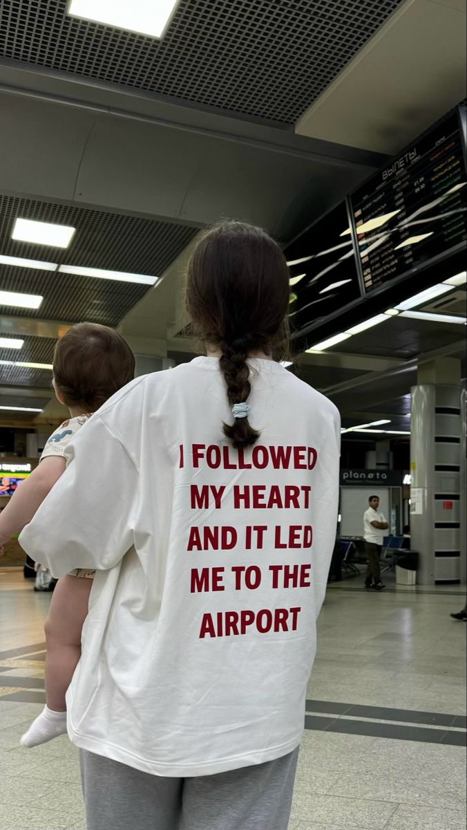 A person standing in an airport terminal wearing a white oversized T-shirt from the back, which features the red text 'I FOLLOWED MY HEART AND IT LED ME TO THE AIRPORT'. The person is holding a baby on their shoulder.