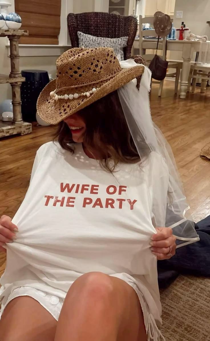 A woman wearing a straw cowboy hat with pearl accents, a white bridal veil, and a white t-shirt with 'WIFE OF THE PARTY' printed in red, sitting in a home interior.