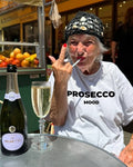 A woman at an outdoor cafe table wearing a white t-shirt with 'PROSECCO MOOD' printed on the front, a black crochet hat, and making a peace sign. A bottle of Prosecco and a filled glass are on the table in front of her.