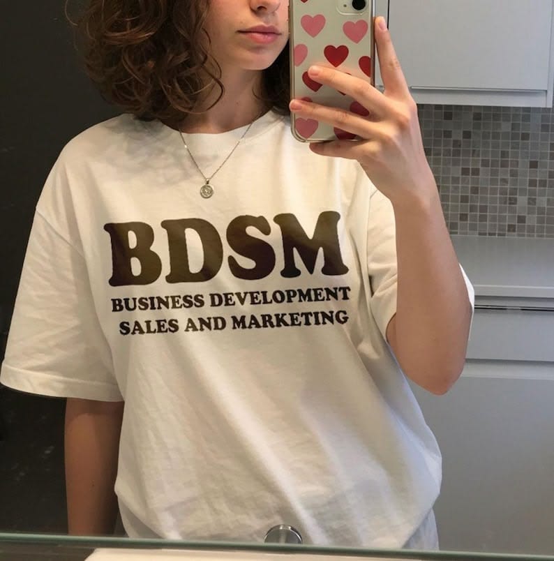 A woman takes a mirror selfie wearing an oversized white t-shirt with the text 'BDSM BUSINESS DEVELOPMENT SALES AND MARKETING' printed in dark brown on the front.