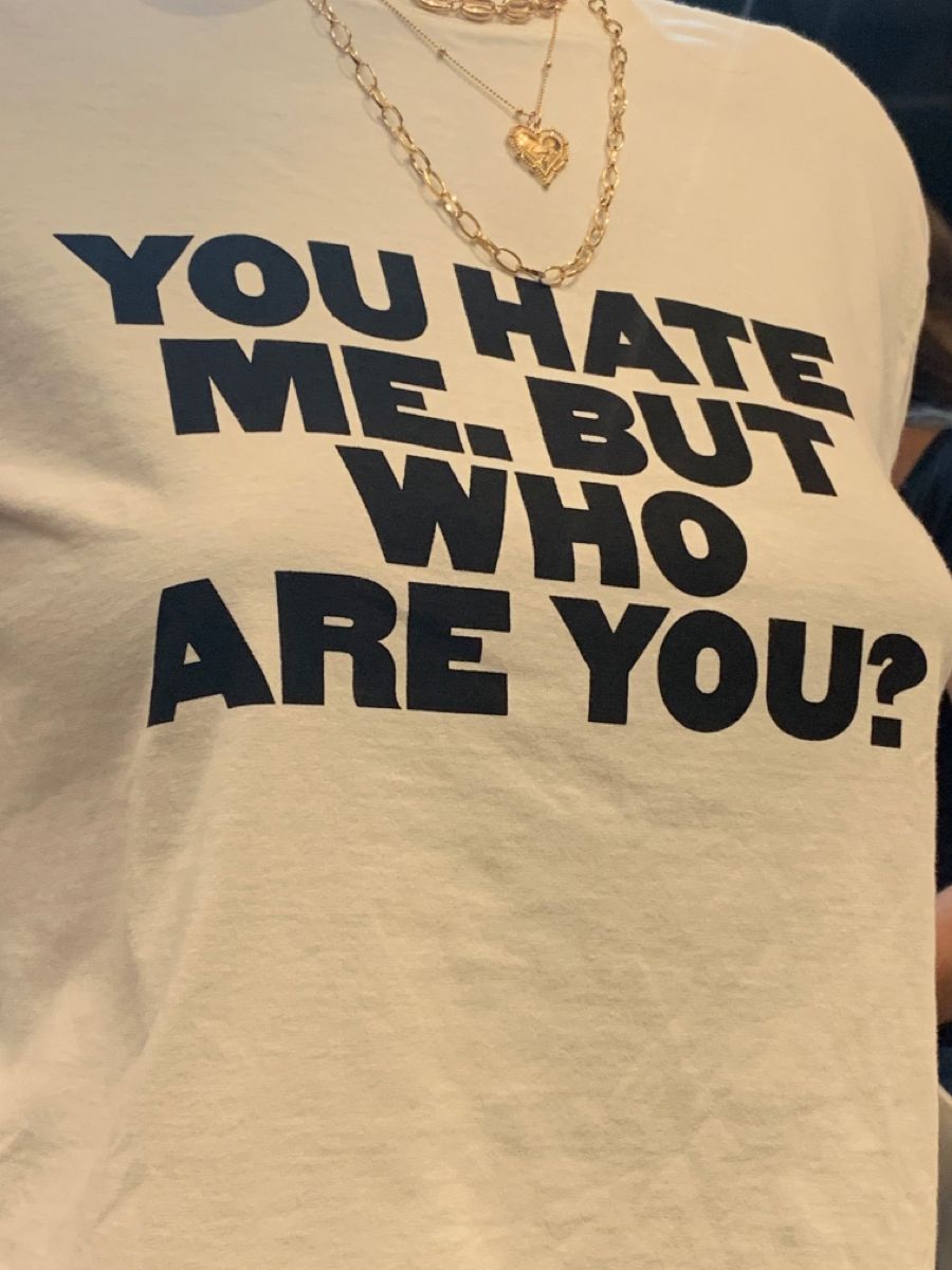 Beige t-shirt with black text 'YOU HATE ME. BUT WHO ARE YOU?' worn by a person.