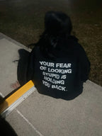 A person viewed from behind wearing a black hoodie with the phrase 'YOUR FEAR OF LOOKING STUPID IS HOLDING YOU BACK.' printed in white text on the back, sitting outdoors on a concrete surface at night.