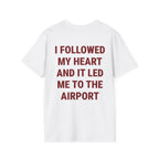 A white short-sleeve t-shirt with bold dark red text on the back that reads 'I FOLLOWED MY HEART AND IT LED ME TO THE AIRPORT', shown against a plain white background.