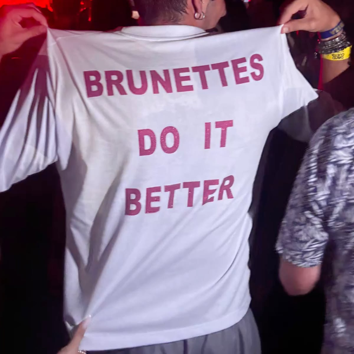 A person wearing a white t-shirt with the phrase 'BRUNETTES DO IT BETTER' printed in pink across the back, held up by hands in a dimly lit setting.