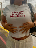 A woman wearing a white short-sleeve crop top T-shirt with the text 'kiss me it's my birthday' printed in red across the chest, posing with her hands on her stomach at what appears to be a train station platform.