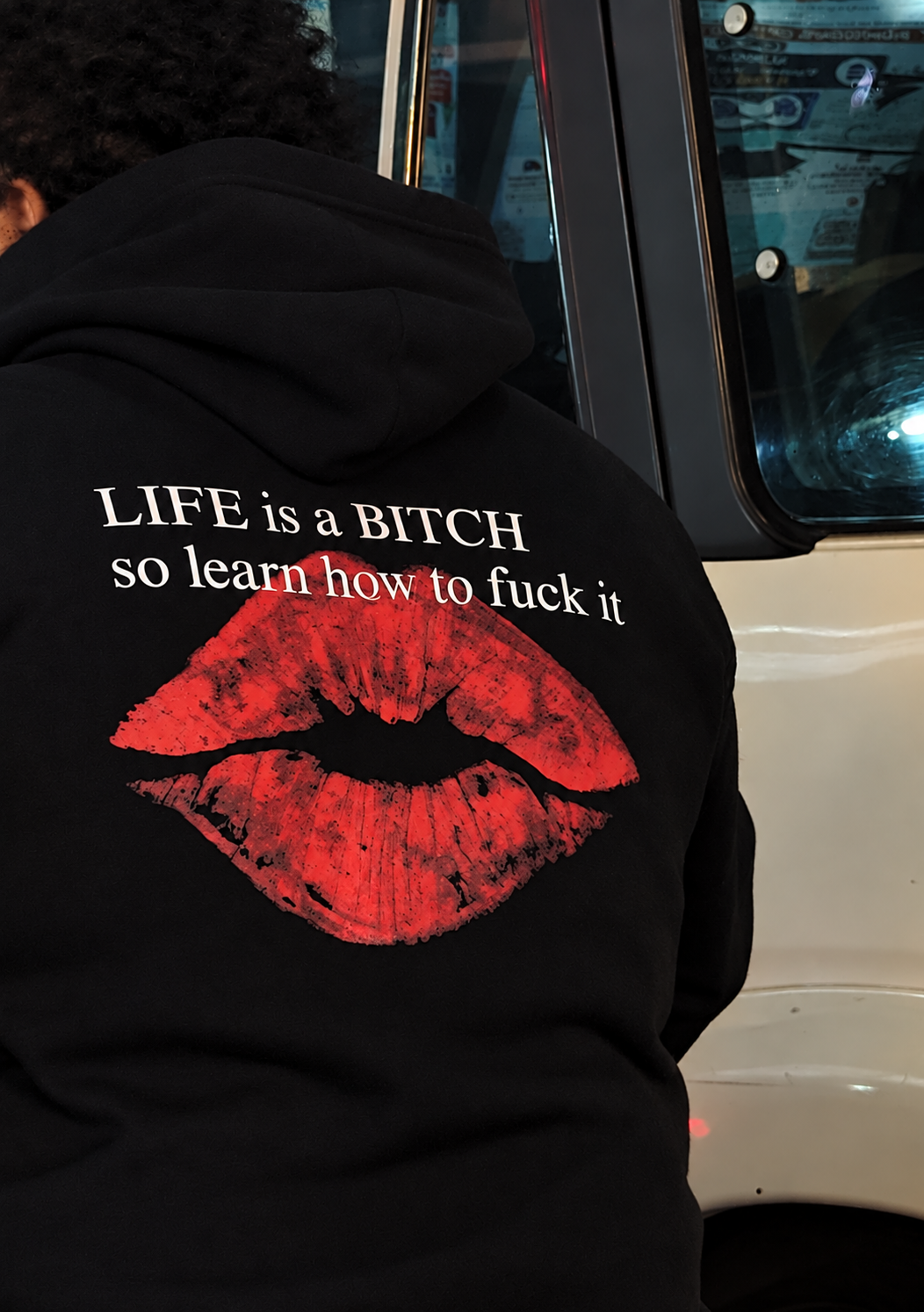 A person wearing a black hoodie from the back, featuring a large red lips graphic and the white text 'LIFE is a BITCH so learn how to fuck it'.