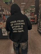 A person standing in a grocery store aisle, viewed from behind, wearing a black hoodie with large white text on the back that reads, 'YOUR FEAR OF LOOKING STUPID IS HOLDING YOU BACK.'.