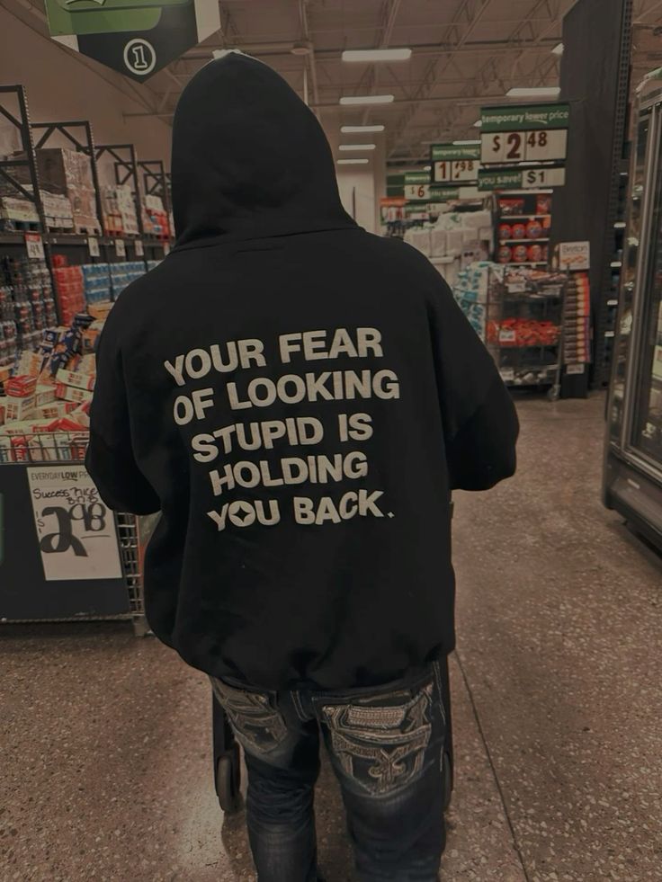 A person standing in a grocery store aisle, viewed from behind, wearing a black hoodie with large white text on the back that reads, 'YOUR FEAR OF LOOKING STUPID IS HOLDING YOU BACK.'.
