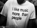 A black and white close-up of a person wearing a white graphic t-shirt with the text 'i like music more than people' printed in black lowercase letters.