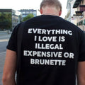 Person wearing a black t-shirt with humorous text on the back in an urban setting. Everything I Love Is Illegal Expensive Or Brunette” Tee