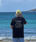 A man standing on a beach with his back to the camera, wearing a black t-shirt with the white text 'WHERE IS MY FUCKING YACHT?' on the back, a yellow cap, and blue patterned swim trunks, looking out at the ocean.