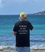 A man standing on a beach with his back to the camera, wearing a black t-shirt with the white text 'WHERE IS MY FUCKING YACHT?' on the back, a yellow cap, and blue patterned swim trunks, looking out at the ocean.