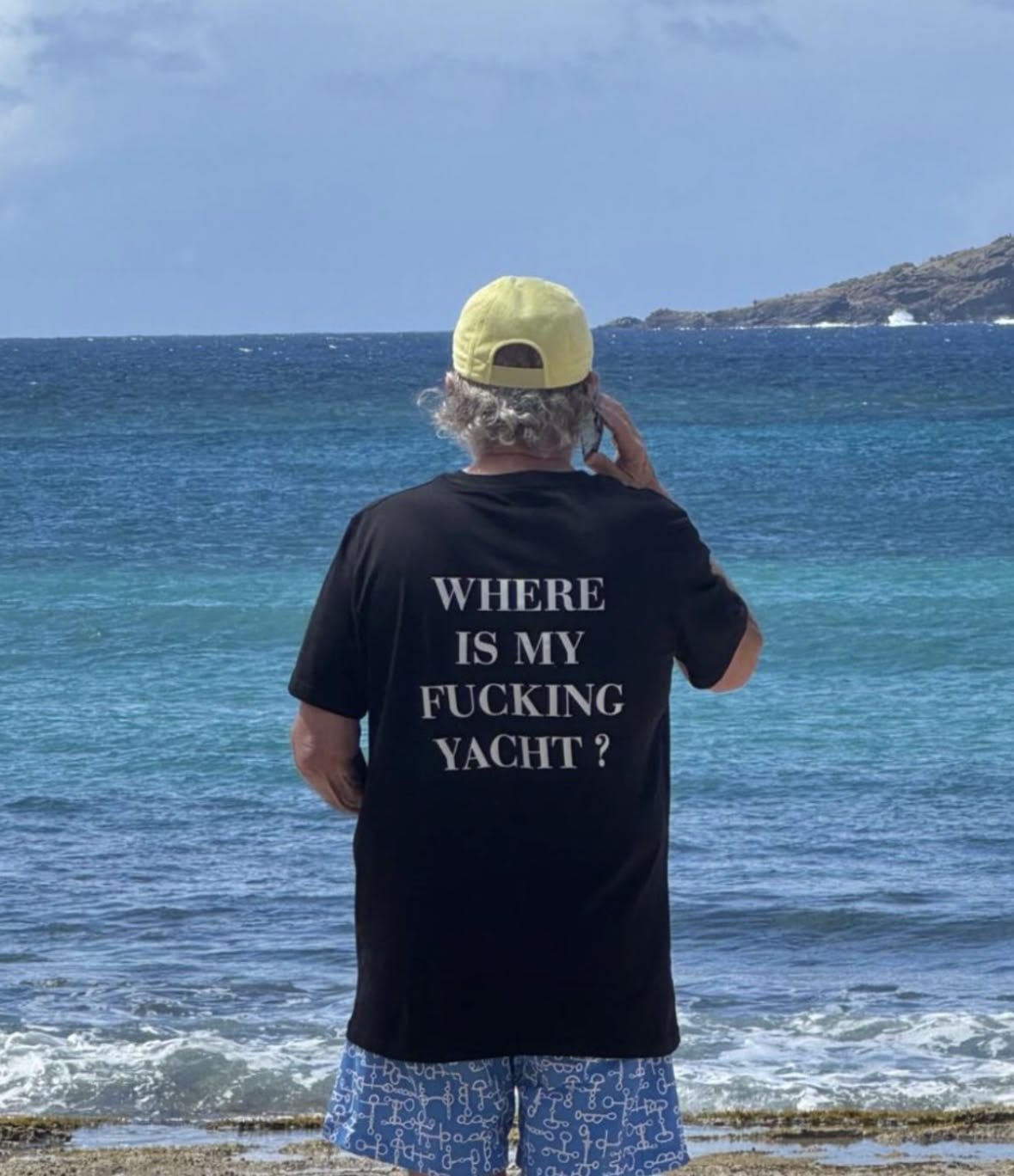 A man standing on a beach with his back to the camera, wearing a black t-shirt with the white text 'WHERE IS MY FUCKING YACHT?' on the back, a yellow cap, and blue patterned swim trunks, looking out at the ocean.